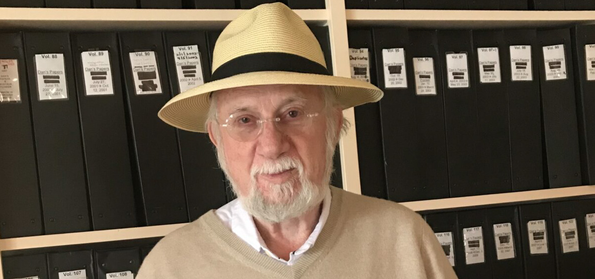 Dan Rattiner: a man wearing a hat in front of black books on a shelves.